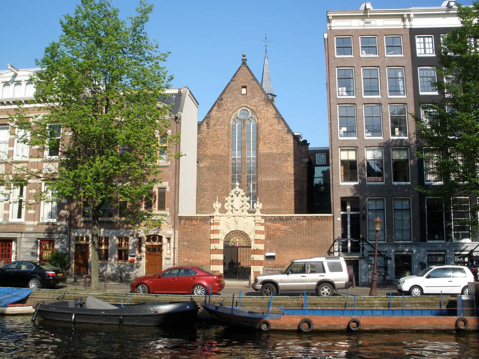 The red-brick Agnietenkapel with its ornate Renaissance gateway on an Amsterdam canal, between tall brick canal houses, with boats moored in the foreground