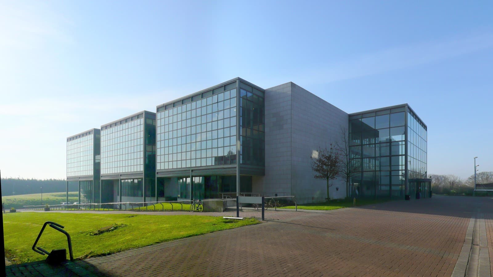 A modern glass-walled library building on Dublin City University's Glasnevin campus, with a brick-paved path and a lawn in front under a clear sky