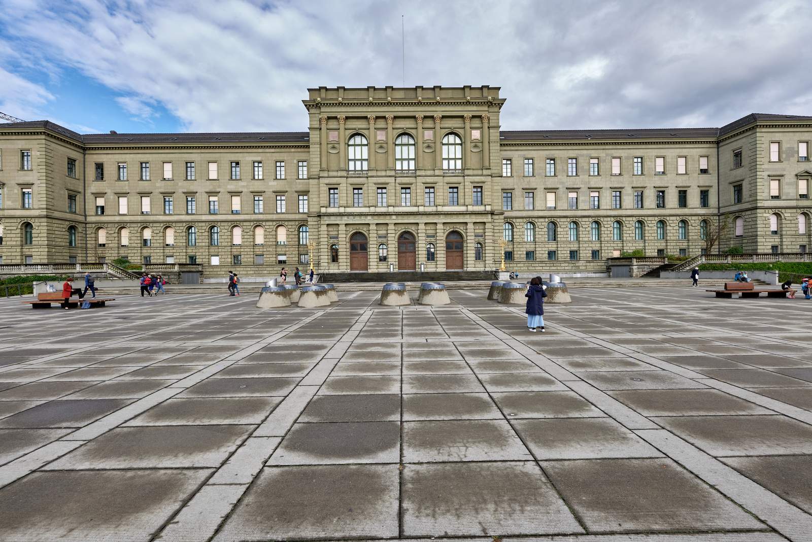 The long neoclassical main building of ETH Zurich seen across the wide paved Polyterrasse under a cloudy sky, with a few people crossing the square