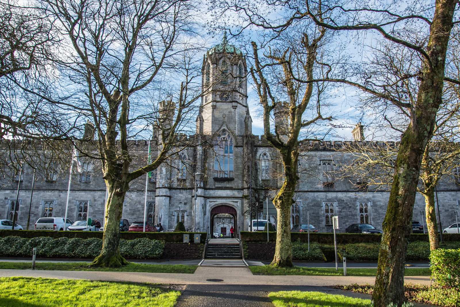 The grey stone Quadrangle building of the University of Galway, with a central clock tower and arched entrance, framed by bare trees and a lawn