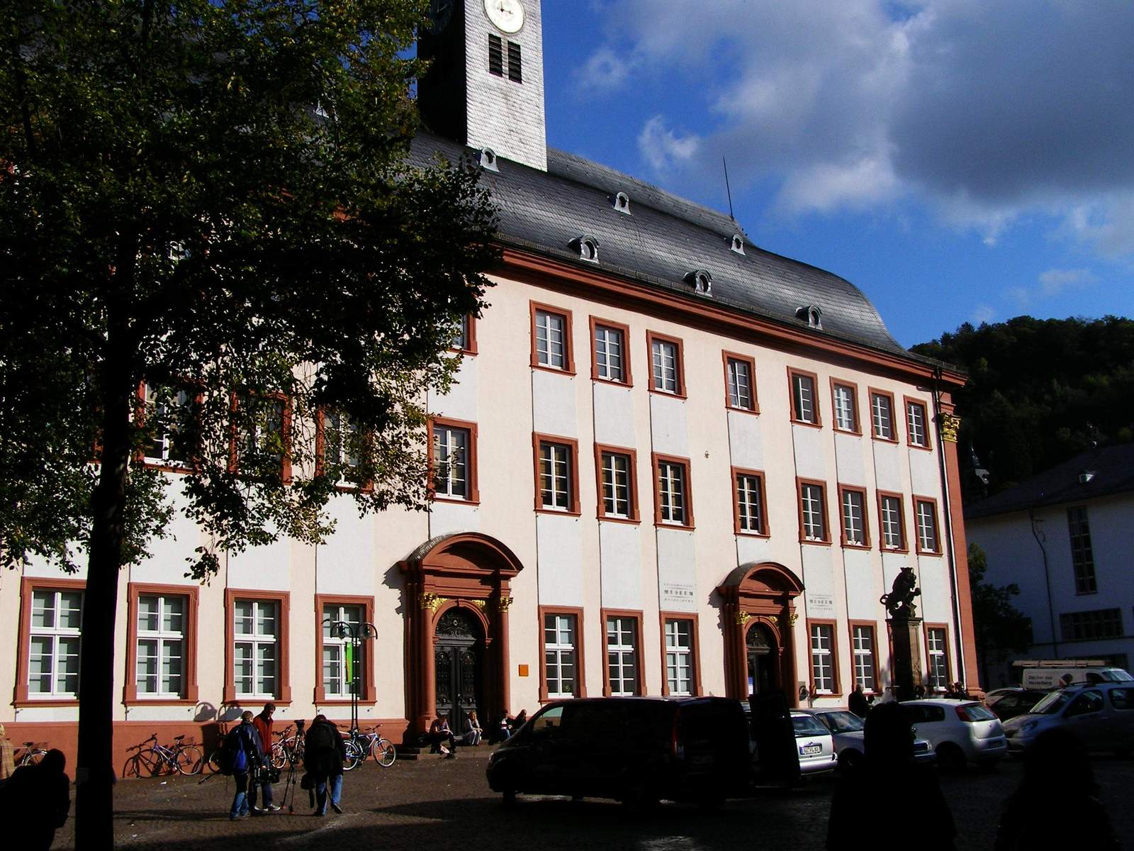 The pale Baroque facade of Heidelberg's Old University building, with red sandstone window frames, two ornate doorways and a small tower, beside a tree on University Square