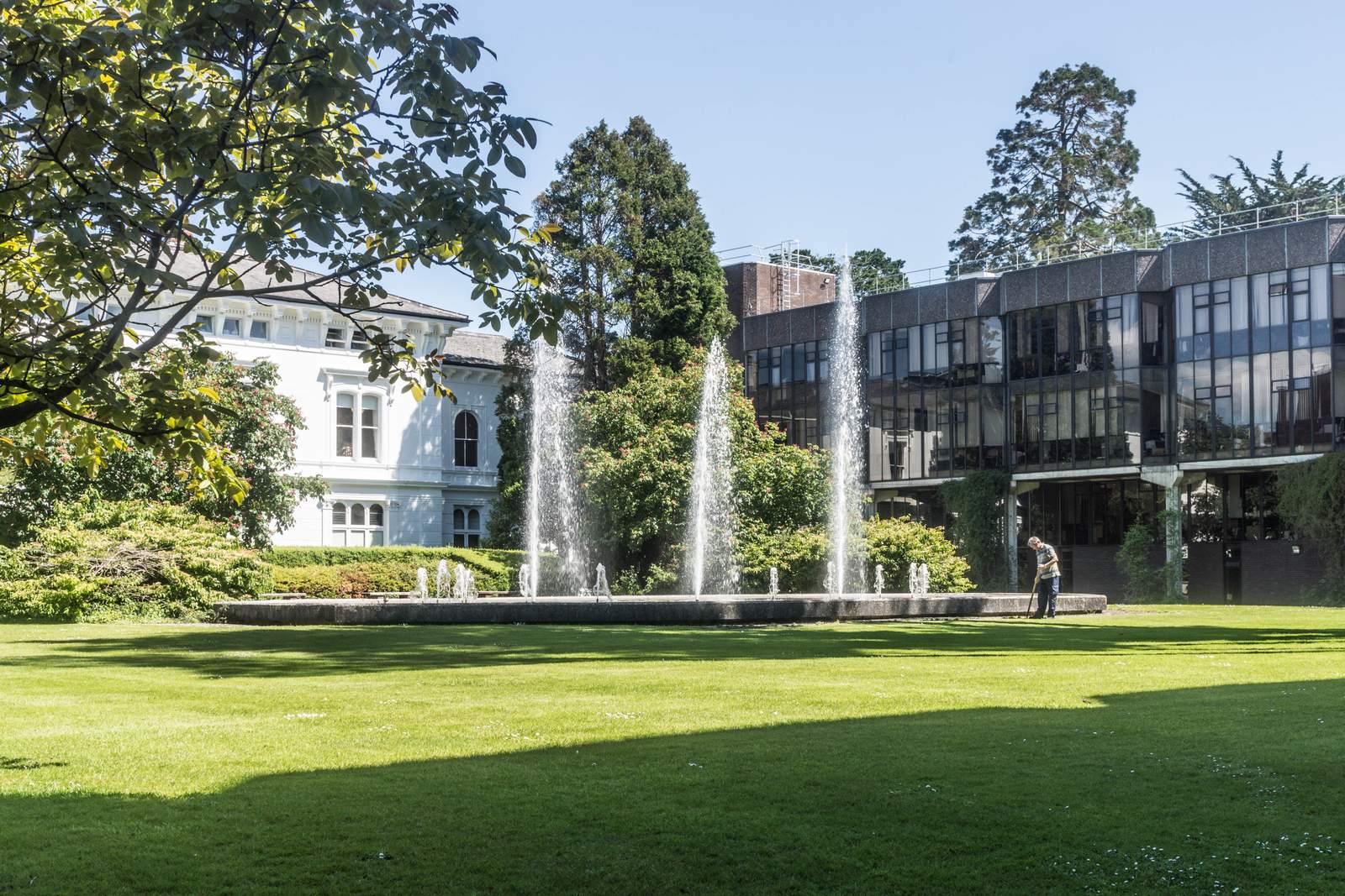 Fountains in front of a white Victorian house and a modern glass-and-steel building on the University of Limerick campus, with a wide sunlit lawn