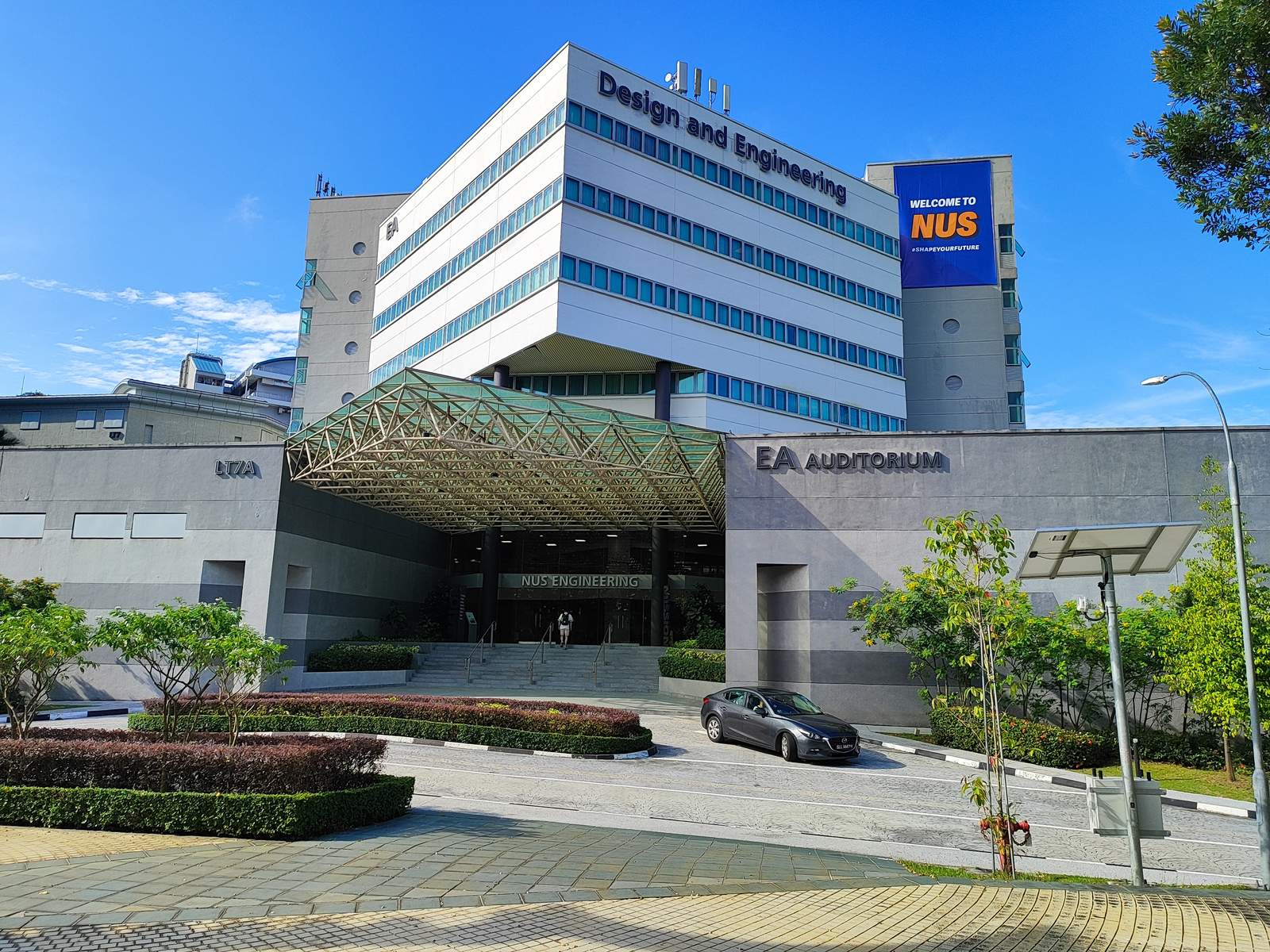 The College of Design and Engineering at the National University of Singapore: a white multi-storey building with a 'Welcome to NUS' banner above a glass-canopied entrance and the EA Auditorium