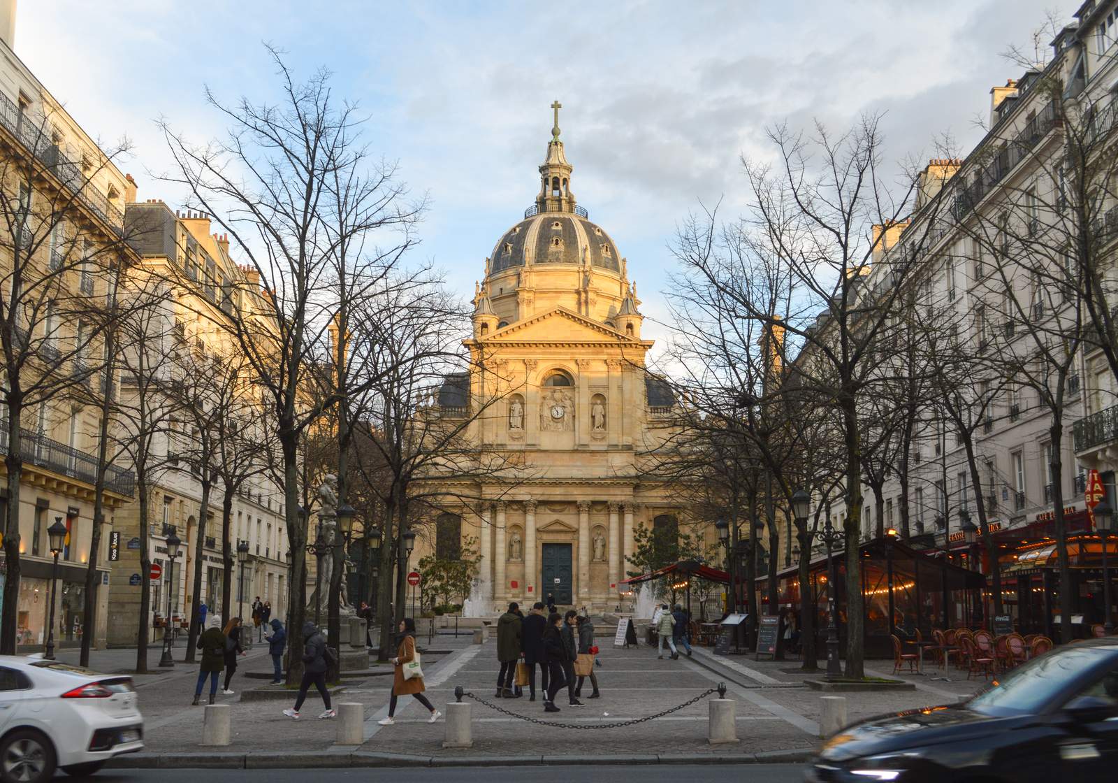 The domed Chapel of the Sorbonne at the end of the Place de la Sorbonne in Paris, lit by late sun, with bare trees, cafe terraces and people walking across the square