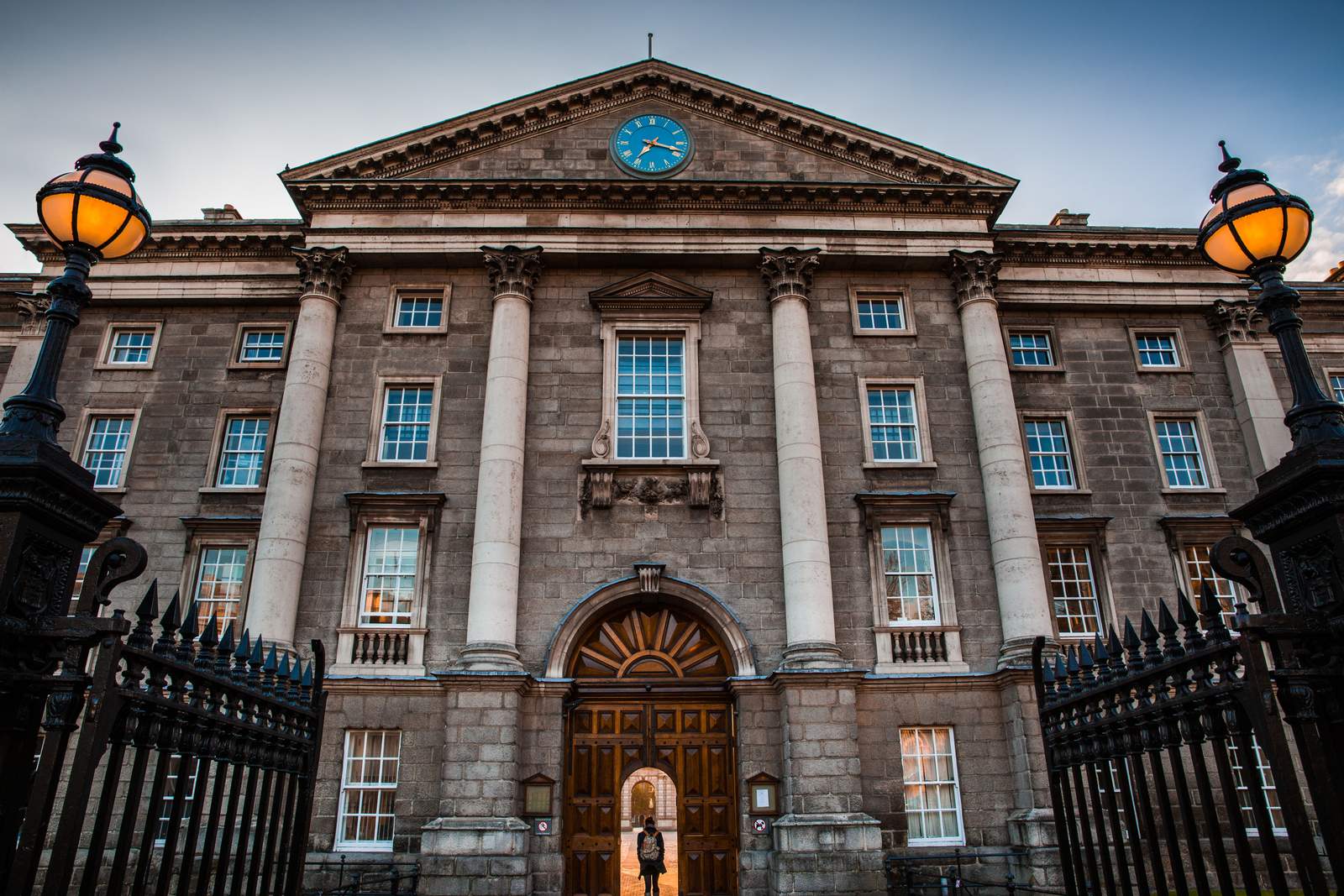 The granite Regent House front of Trinity College Dublin on College Green, with Corinthian columns, a clock in the pediment and a student in the arched gateway between black iron gates and lamps