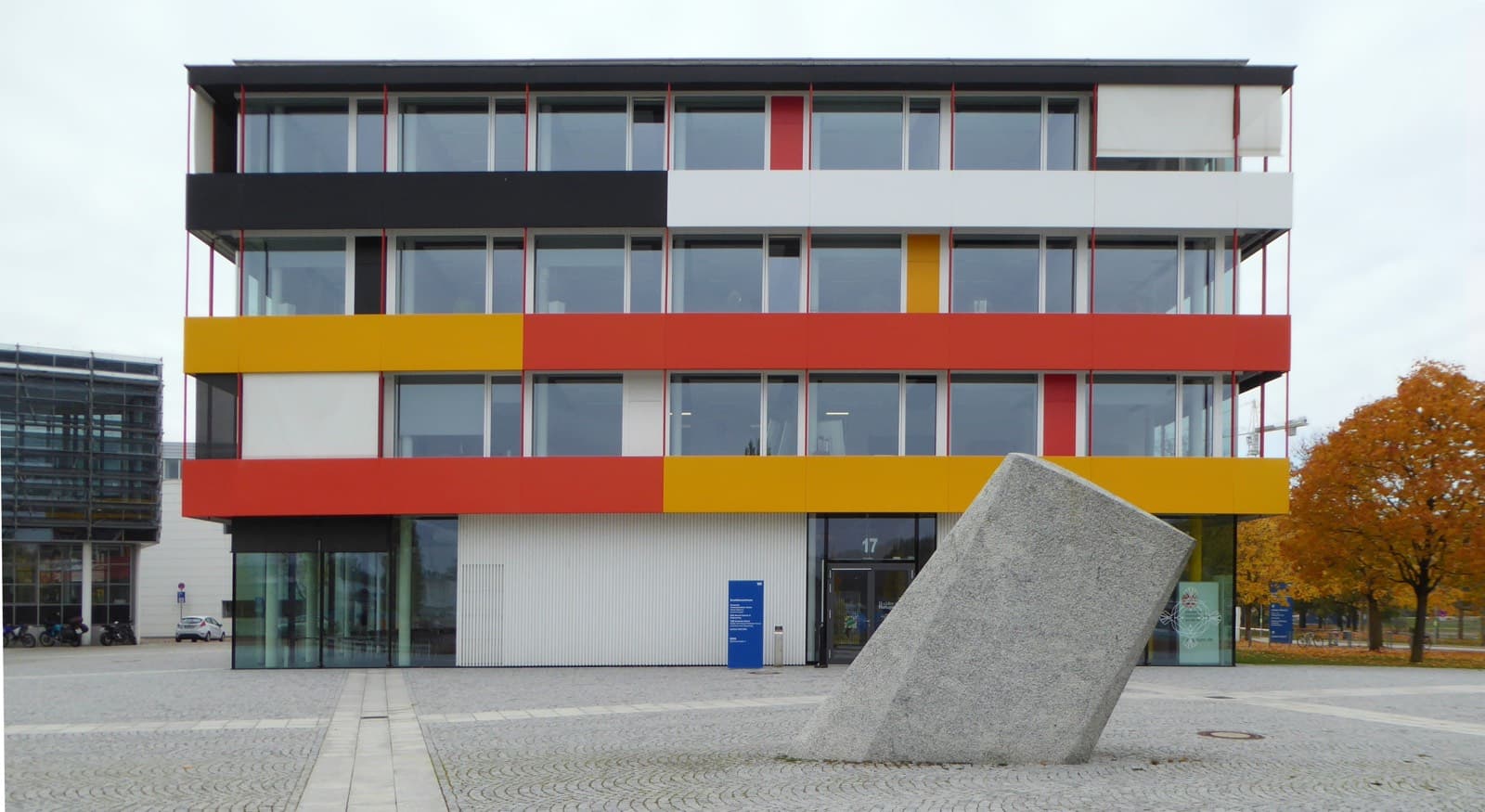 A modern four-storey building with red, yellow, white and black facade panels on TUM's Garching campus, with a large tilted granite block in the paved square in front