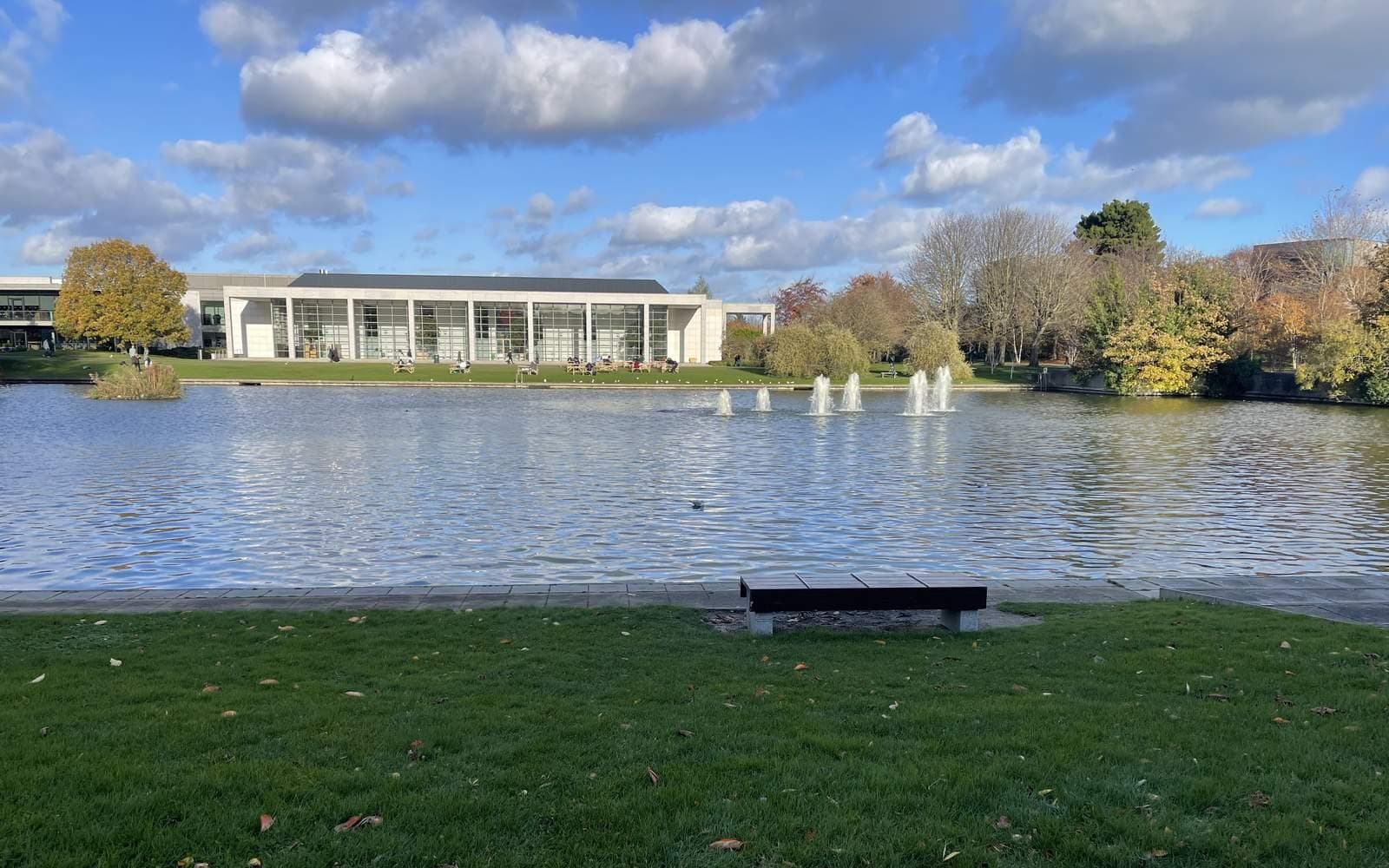 A lake with fountains on UCD's Belfield campus, with a long low glass-fronted building and autumn trees on the far bank under a blue sky